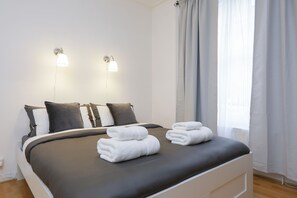 A neatly made bed with white towels, a dark grey bedspread, and two wall-mounted lights.