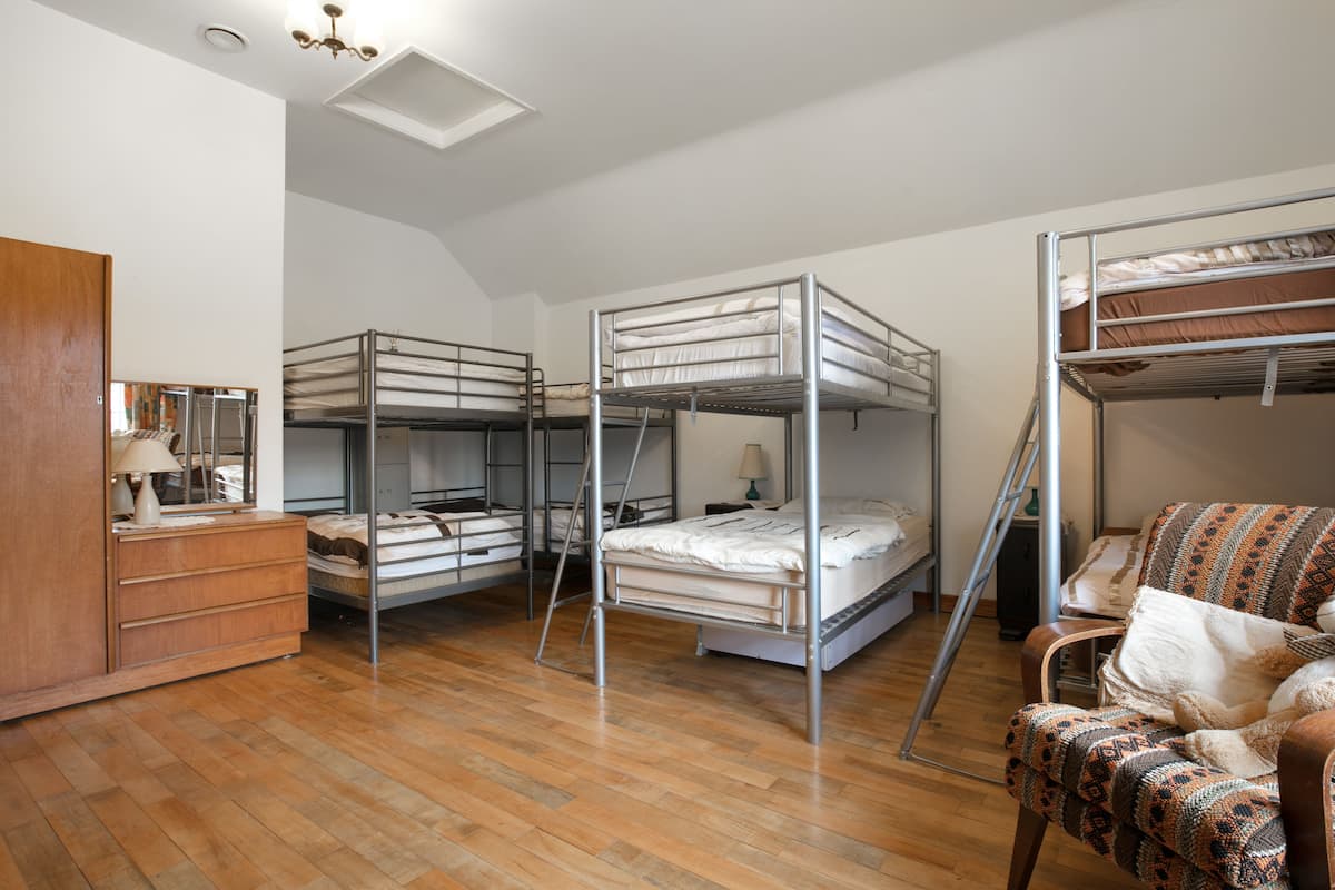 A dormitory room with bunk beds, a wooden dresser, a chair, and a desk with a lamp.