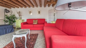 Ein Wohnzimmer mit einem roten Sofa, einer blauen Bank, einem weißen Couchtisch, einem gemusterten Teppich und einer Decke mit freiliegenden Holzbalken.