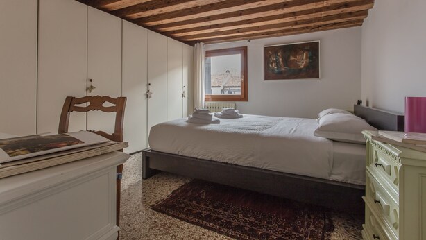 Ein Schlafzimmer mit Holzbalkendecke, einem Bett mit weißer Bettwäsche, einem Fenster mit Blick und einem weißen Kleiderschrank.