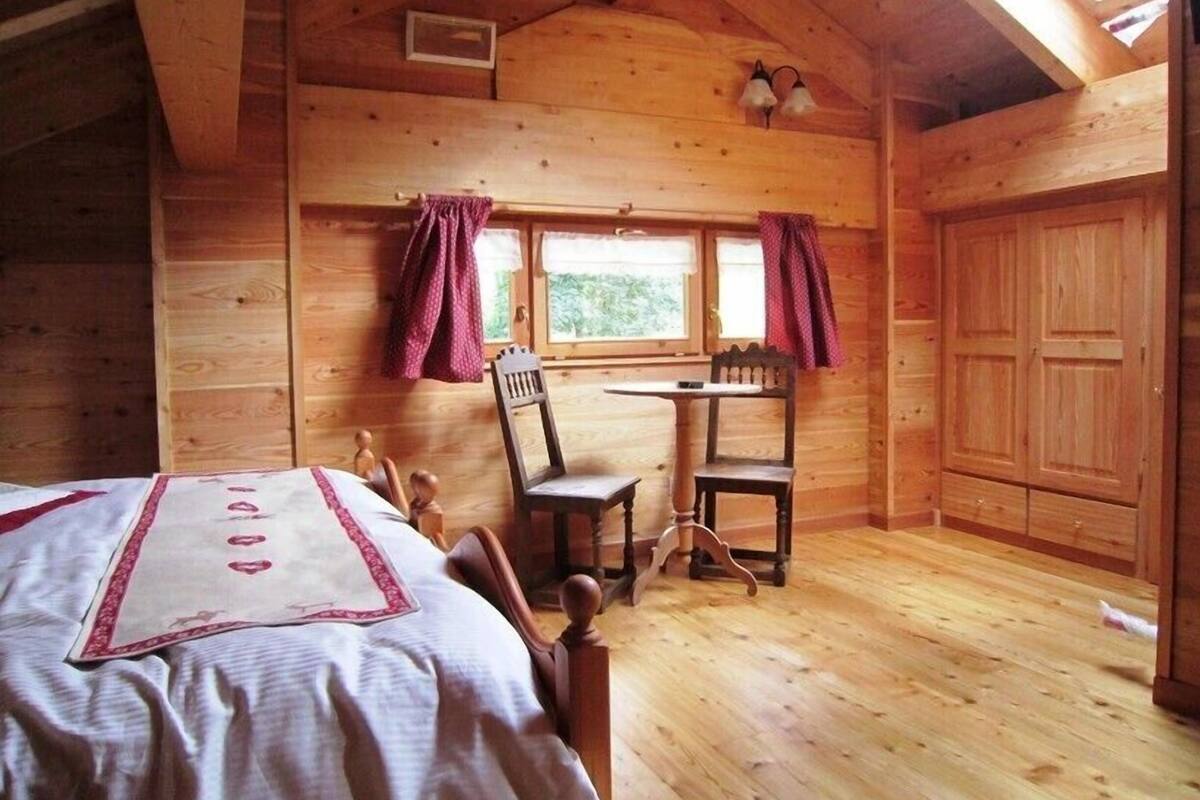 A cozy wooden cabin room with a bed, a table with chairs, a window with curtains, and a wardrobe.