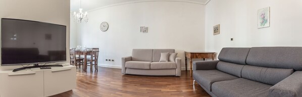 A living room with a flat-screen TV, a grey sofa, a beige sofa, a dining table with chairs, and a chandelier. - Venezia - San Pantalon 3700 (Venice)