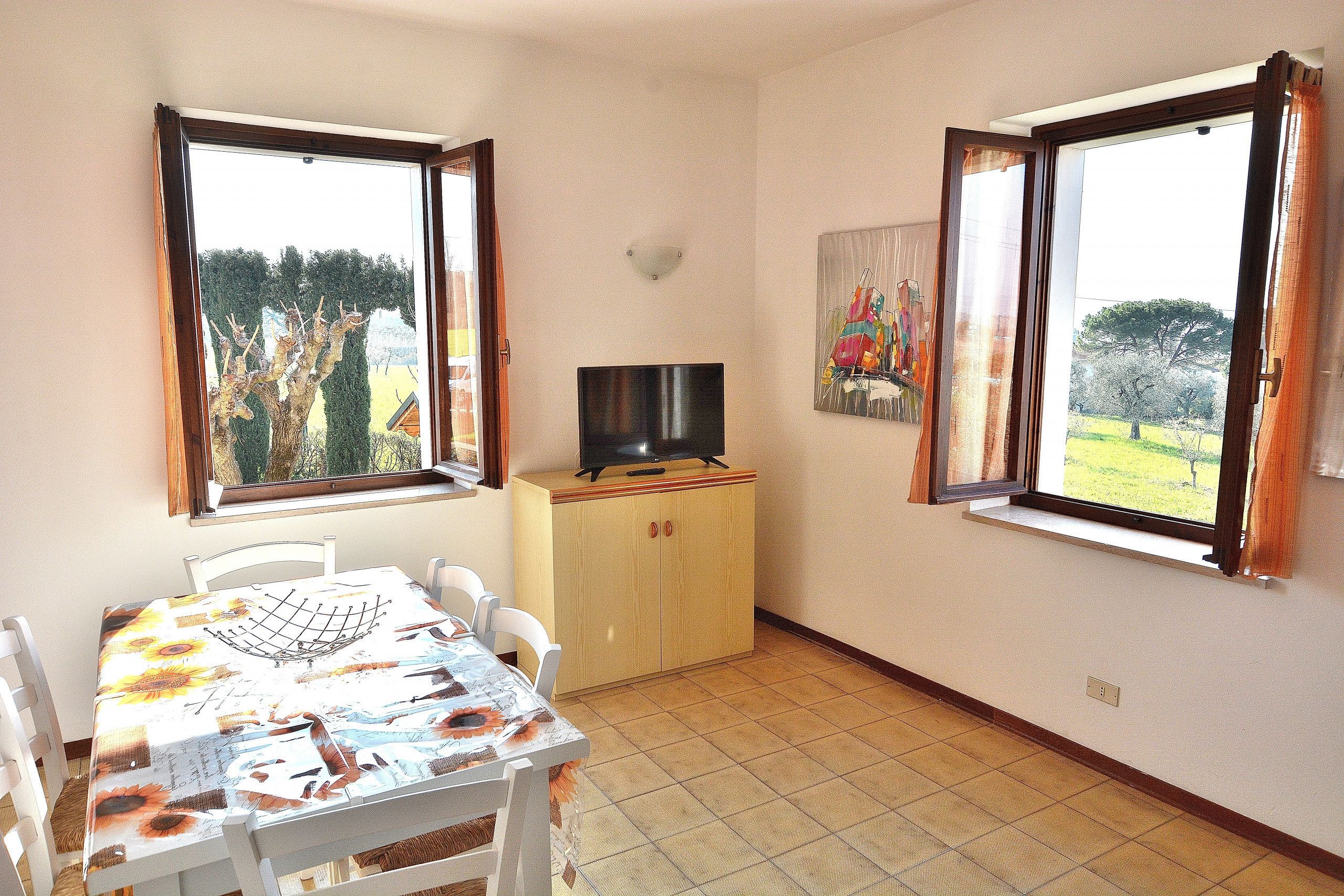 A dining area with a table set for four, a television on a wooden cabinet, and two windows with curtains.