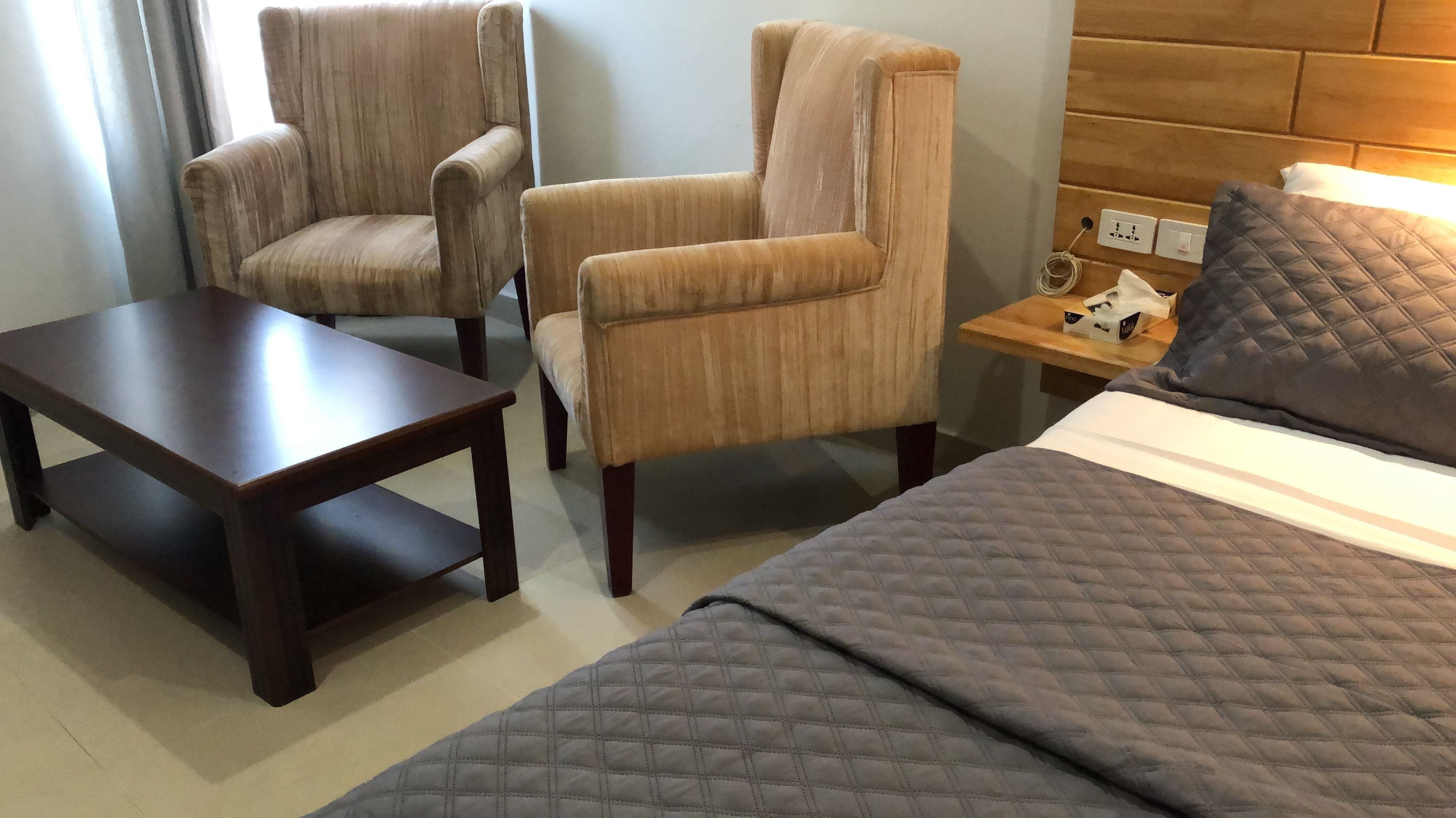 A hotel room with a bed, two armchairs, a small wooden table, and a nightstand with a lamp.