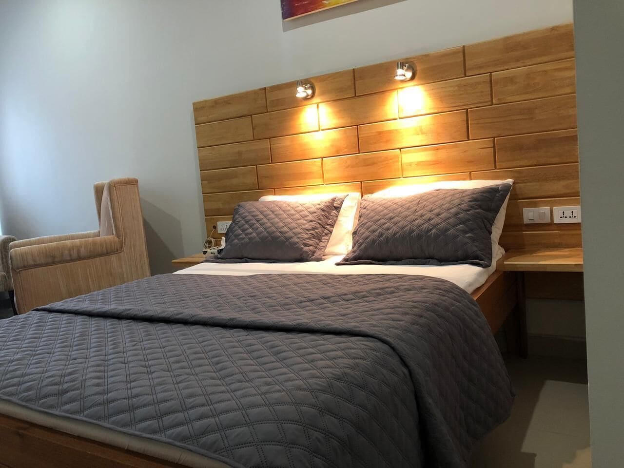 A neatly made bed with a quilted bedspread and two pillows, a wooden headboard with integrated lighting, and a bedside table with a lamp.