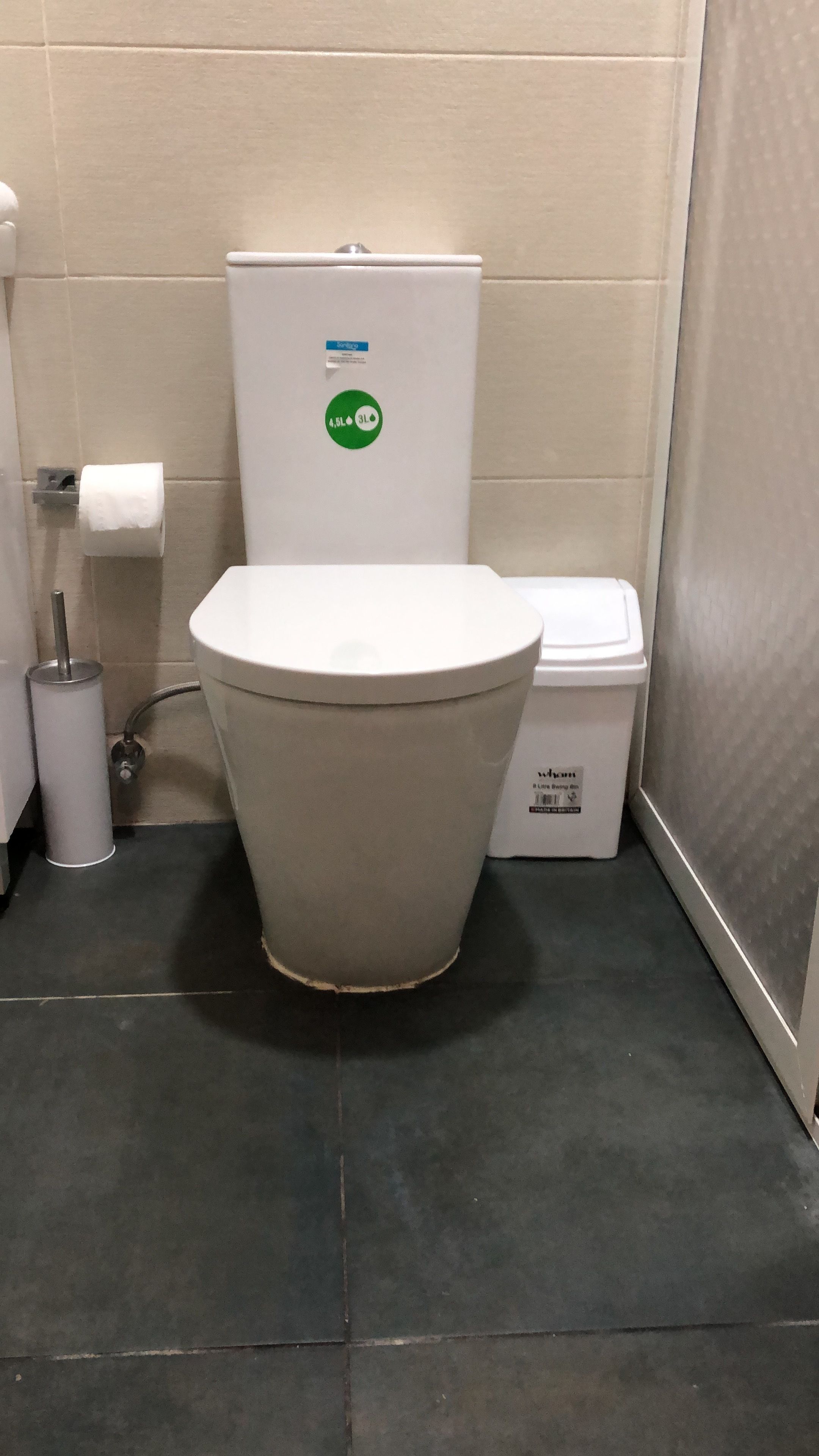 Des toilettes blanches ornées d’un autocollant vert, un porte-rouleau de papier toilette et une poubelle dans une salle de bain dont les murs et le sol sont recouverts de carreaux.
