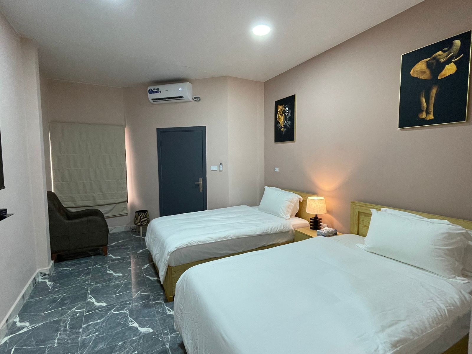 A hotel room with two single beds, a chair, an air conditioning unit, a door, and a wall-mounted artwork.
