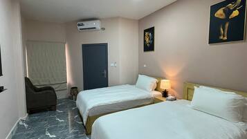 A hotel room with two single beds, a chair, an air conditioning unit, a door, and a wall-mounted artwork.