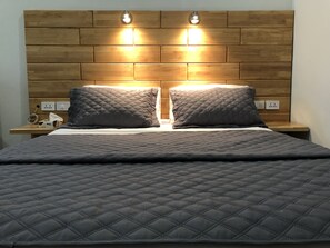 Una cama con colcha gris acolchada, dos mesitas de noche con lámparas y un cabecero de madera con dos luces empotradas.