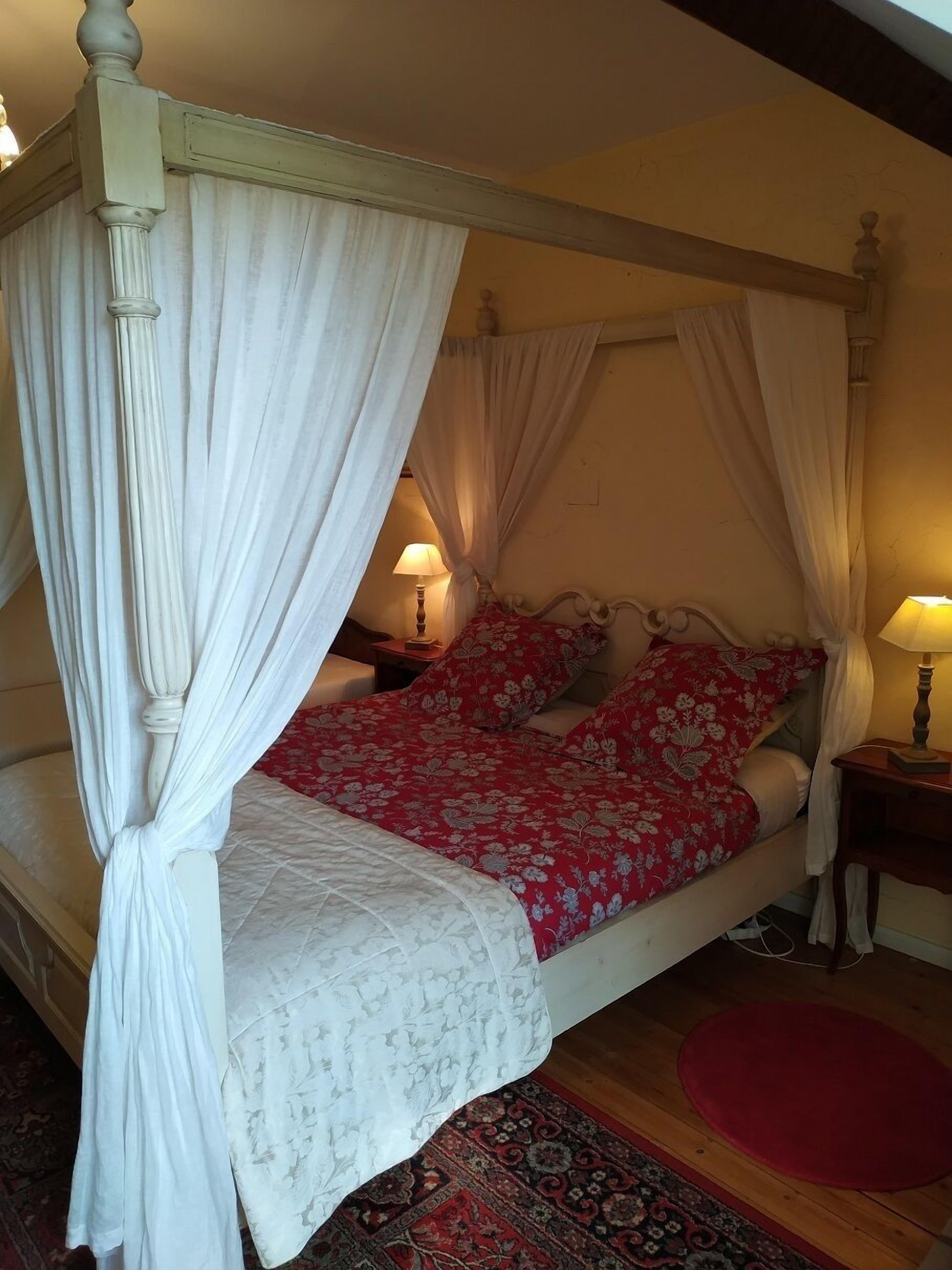 Een hemelbed met witte gordijnen, een rode bloemenovertrek en een witte quilt.