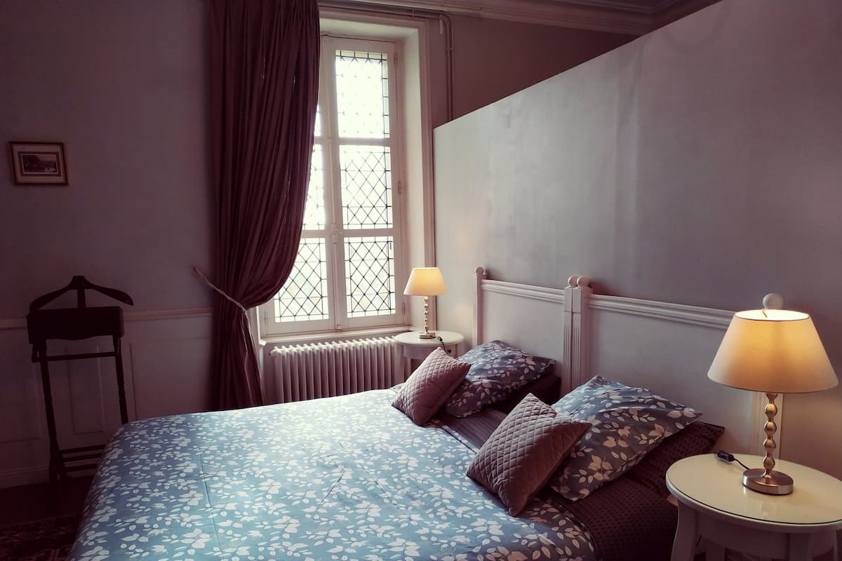 A bedroom with a bed, two bedside lamps, a radiator, a window with curtains, and a chair.