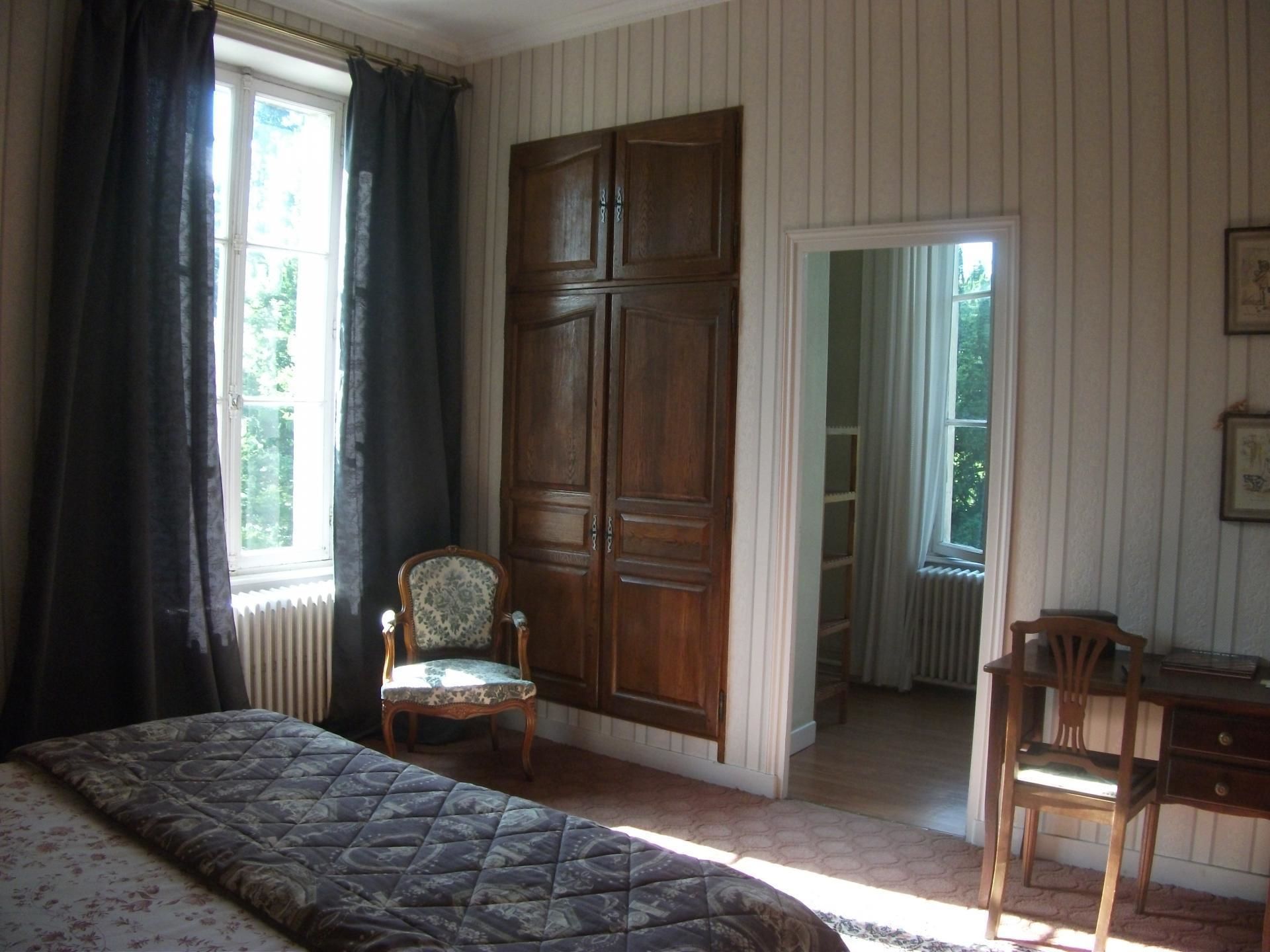 Un dormitorio con cama, dos sillas, un armario de madera y una ventana con cortinas.