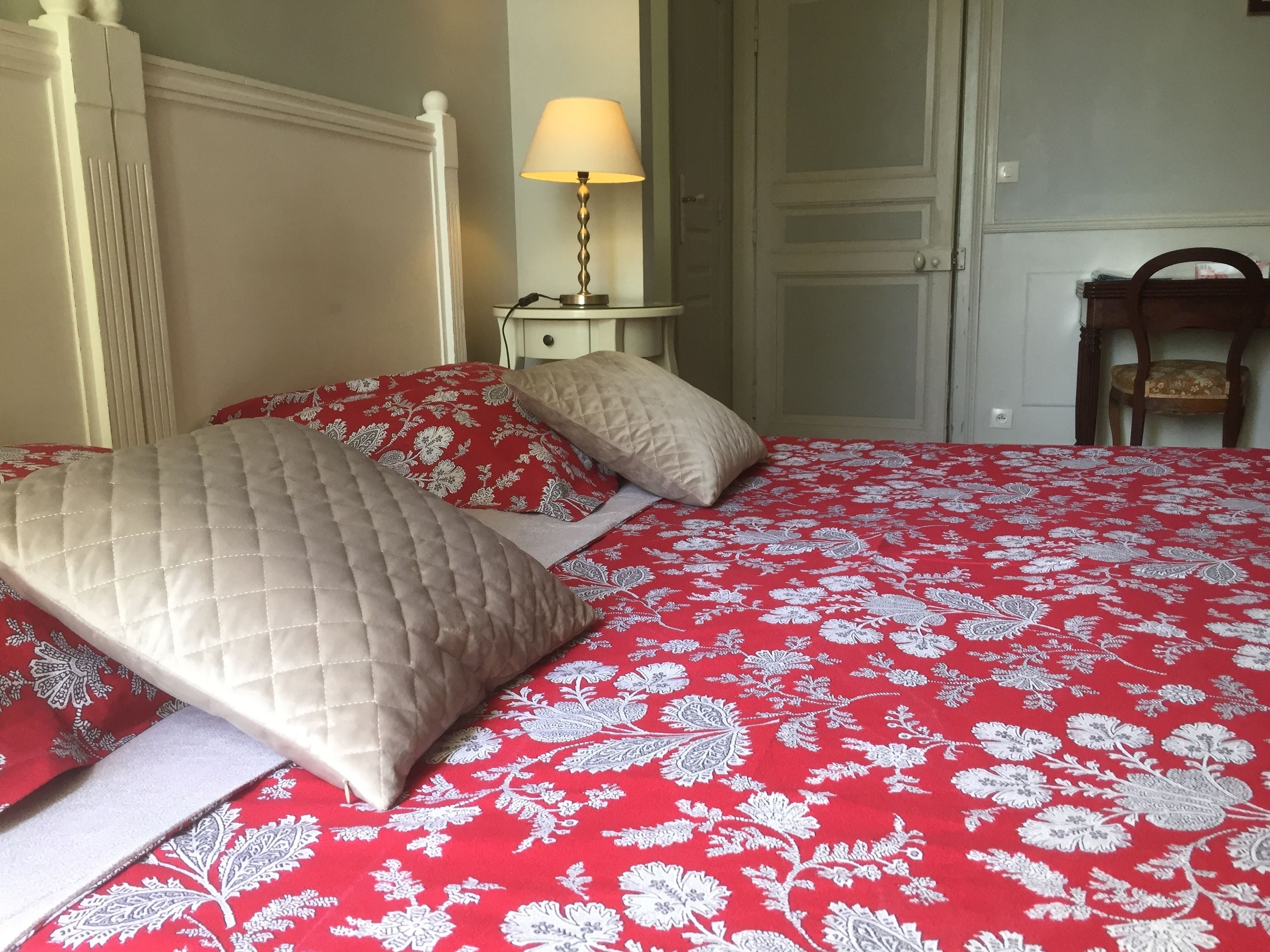 Una cama bien hecha con una colcha roja estampada con flores y dos almohadas, una lámpara de noche y una silla de madera en una habitación con una puerta y una pared blanca.