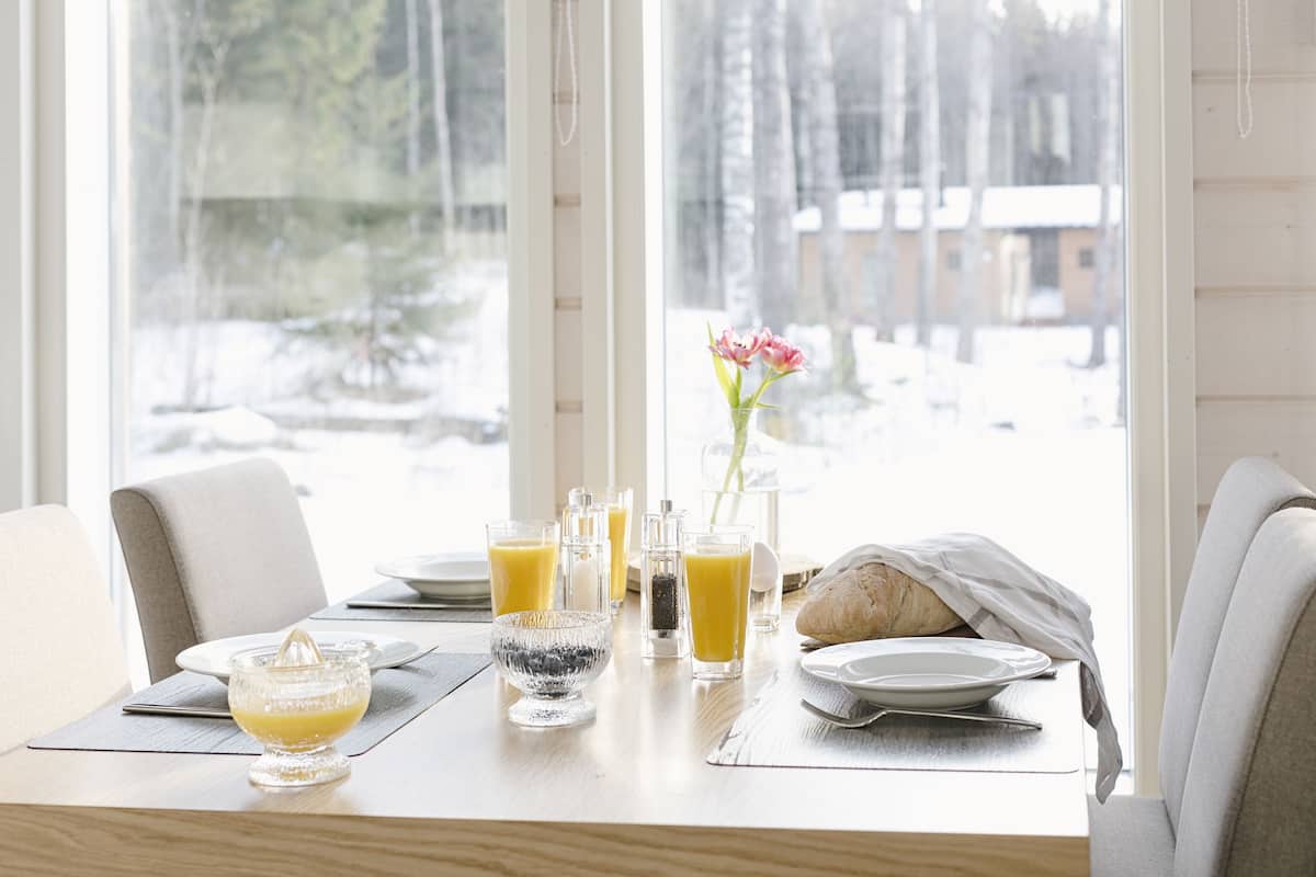 A breakfast table with a glass of orange juice, a glass of water, a bread roll, and a vase with flowers.