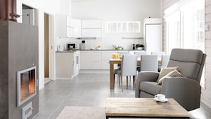 A modern kitchen with white cabinets, a dining area with a table and chairs, and a gray armchair. - Wooddream (Taipalsaari)
