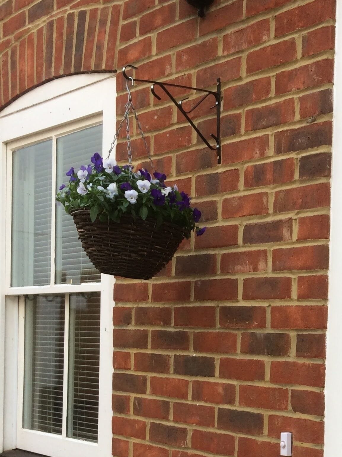 Un panier suspendu rempli de fleurs violettes et blanches, placé devant un mur de briques.