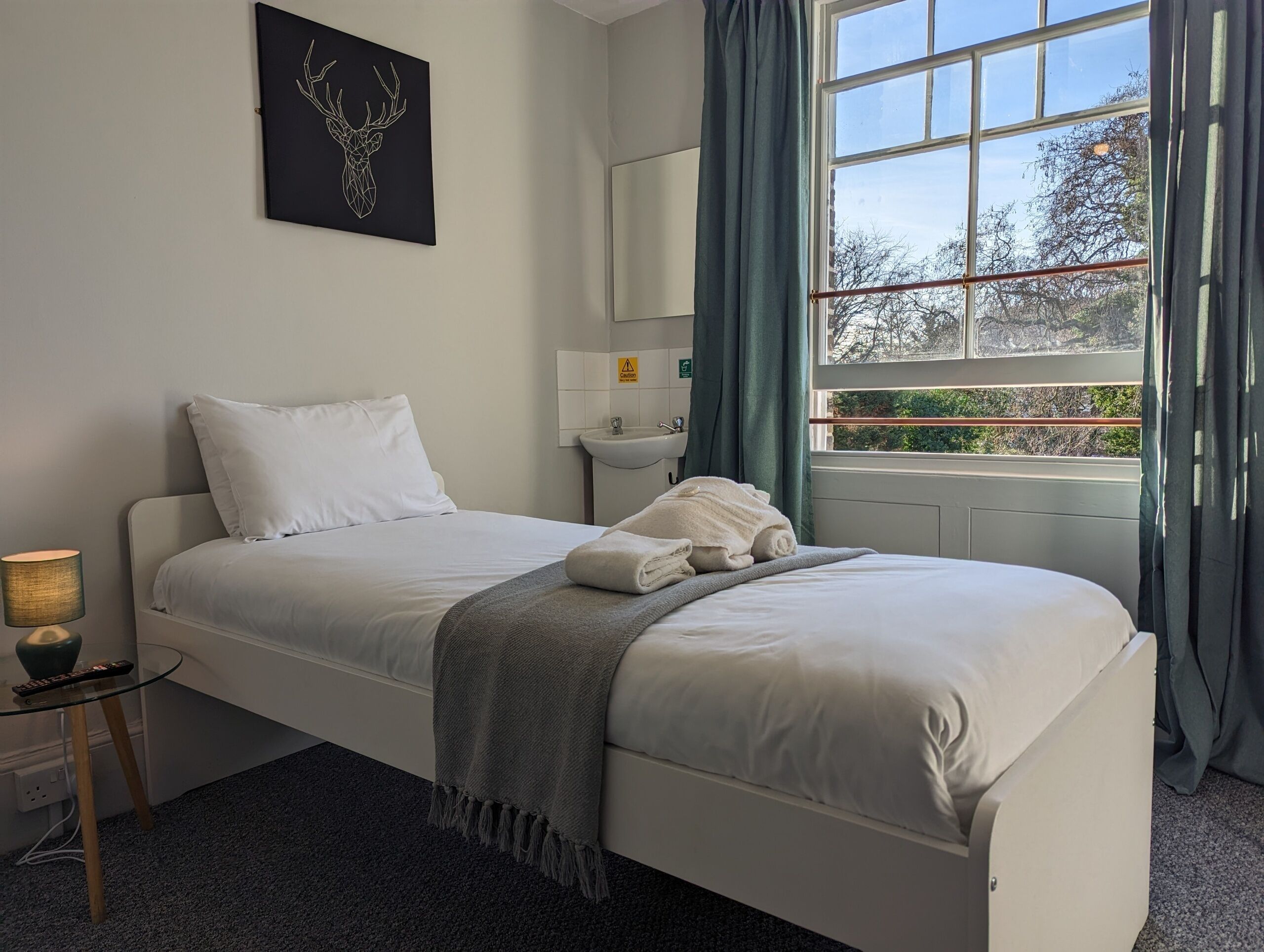 A neatly made bed with white linens, a gray throw blanket, and a folded towel on the side. A small bedside table with a lamp and remote control is visible. A wall-mounted mirror and a window with curtains are also present.
