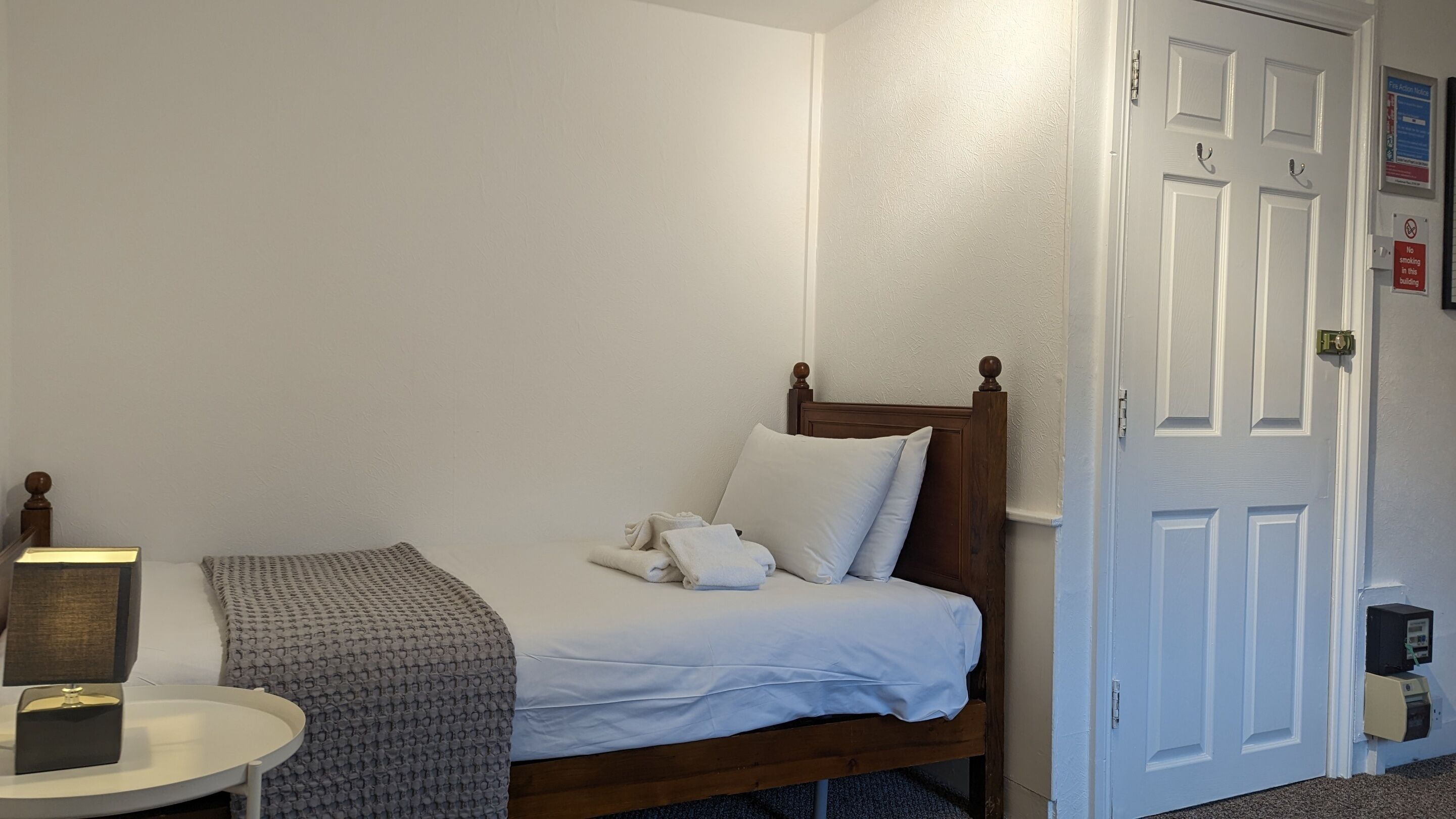 A single bed with a wooden headboard, white bedding, and a bedside table with a lamp and a book.