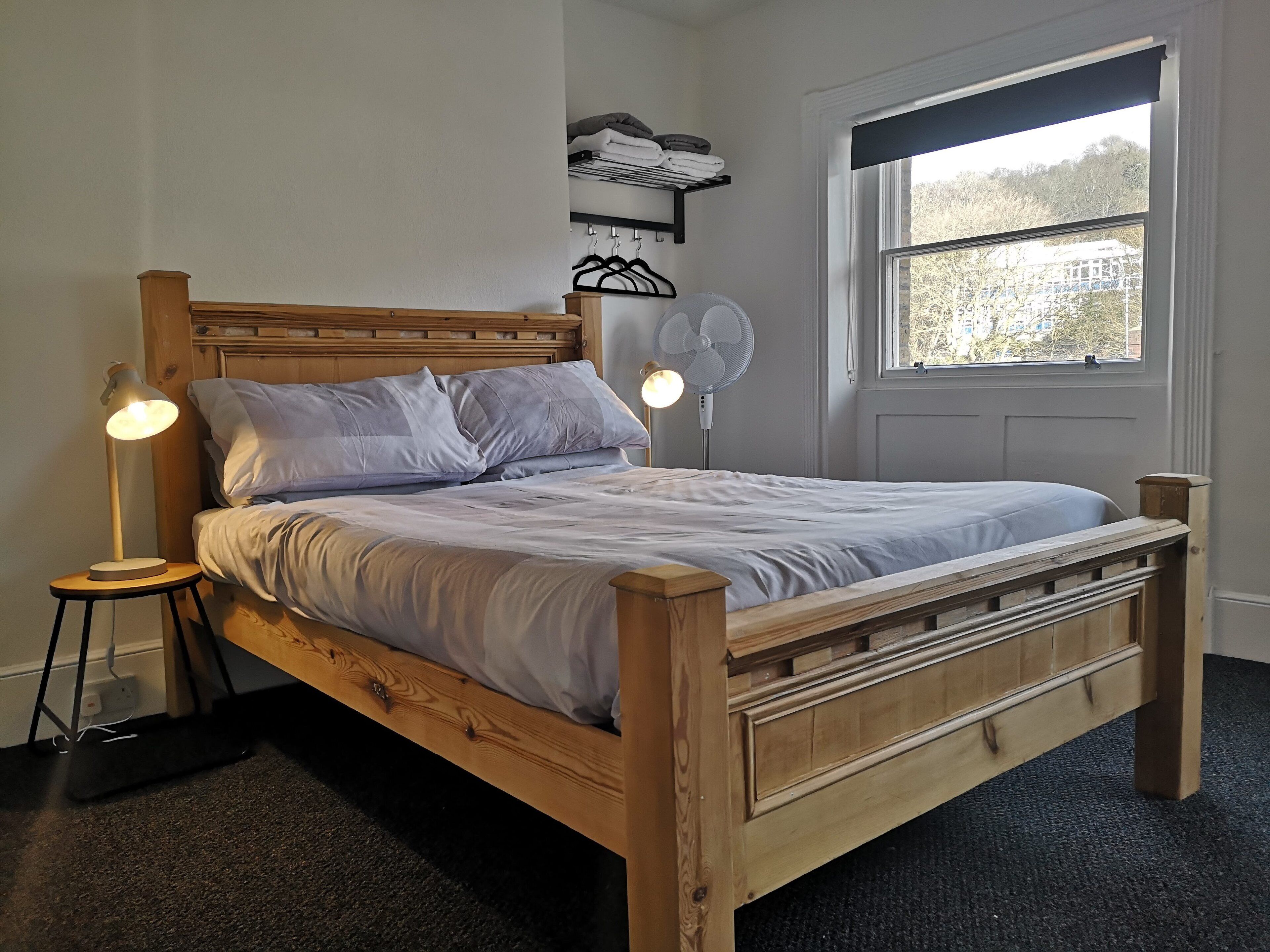 Una cama de madera con cabecera, dos lámparas de noche, un ventilador en la pared y una ventana con vistas a árboles y edificios.