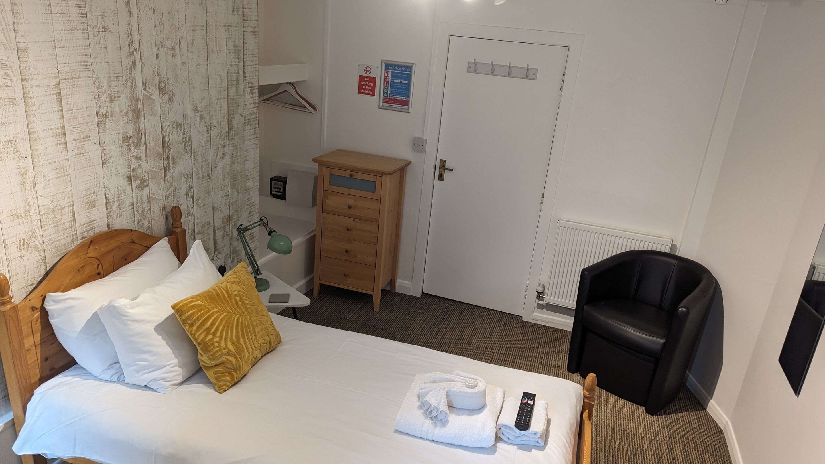 Une chambre d’hôtel avec un lit, une commode en bois, une chaise noire, un bureau avec une lampe et une porte avec une pancarte.