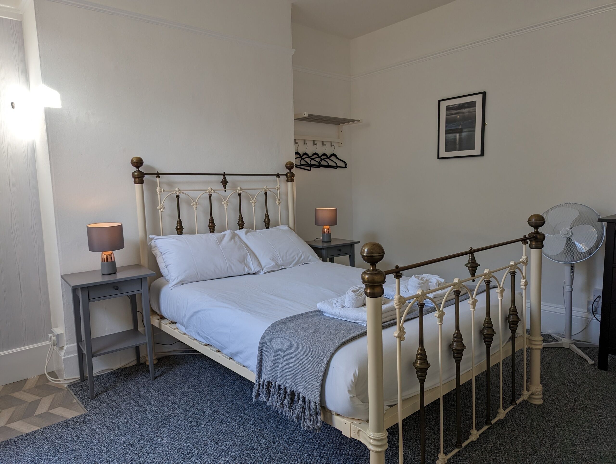 Uma cama bem arrumada com lençóis brancos, duas mesas de cabeceira com luminárias, um quadro pendurado na parede e um ventilador de chão.