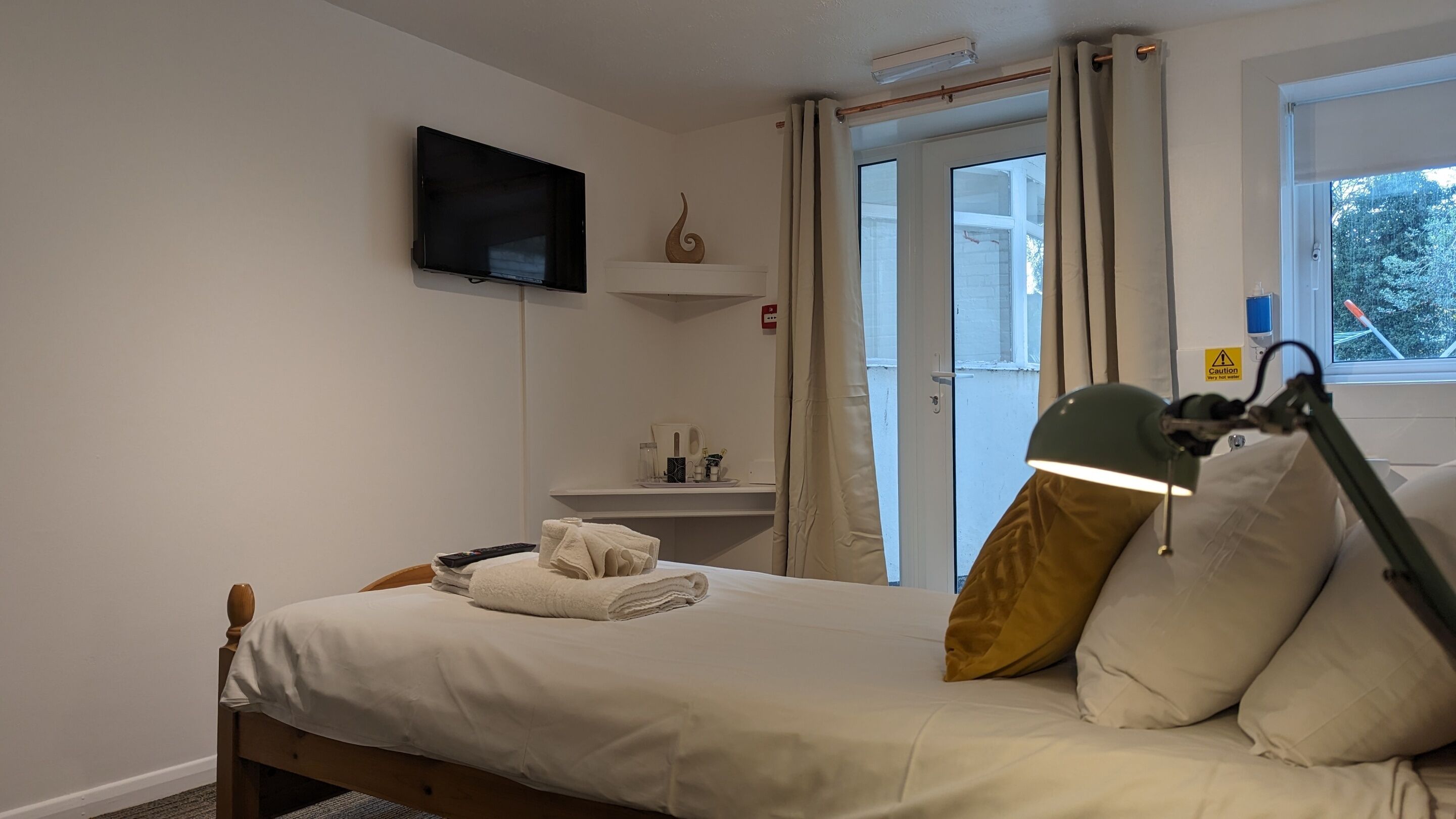 A bedroom with a bed, a television mounted on the wall, a bedside table with a lamp, and a window with curtains.