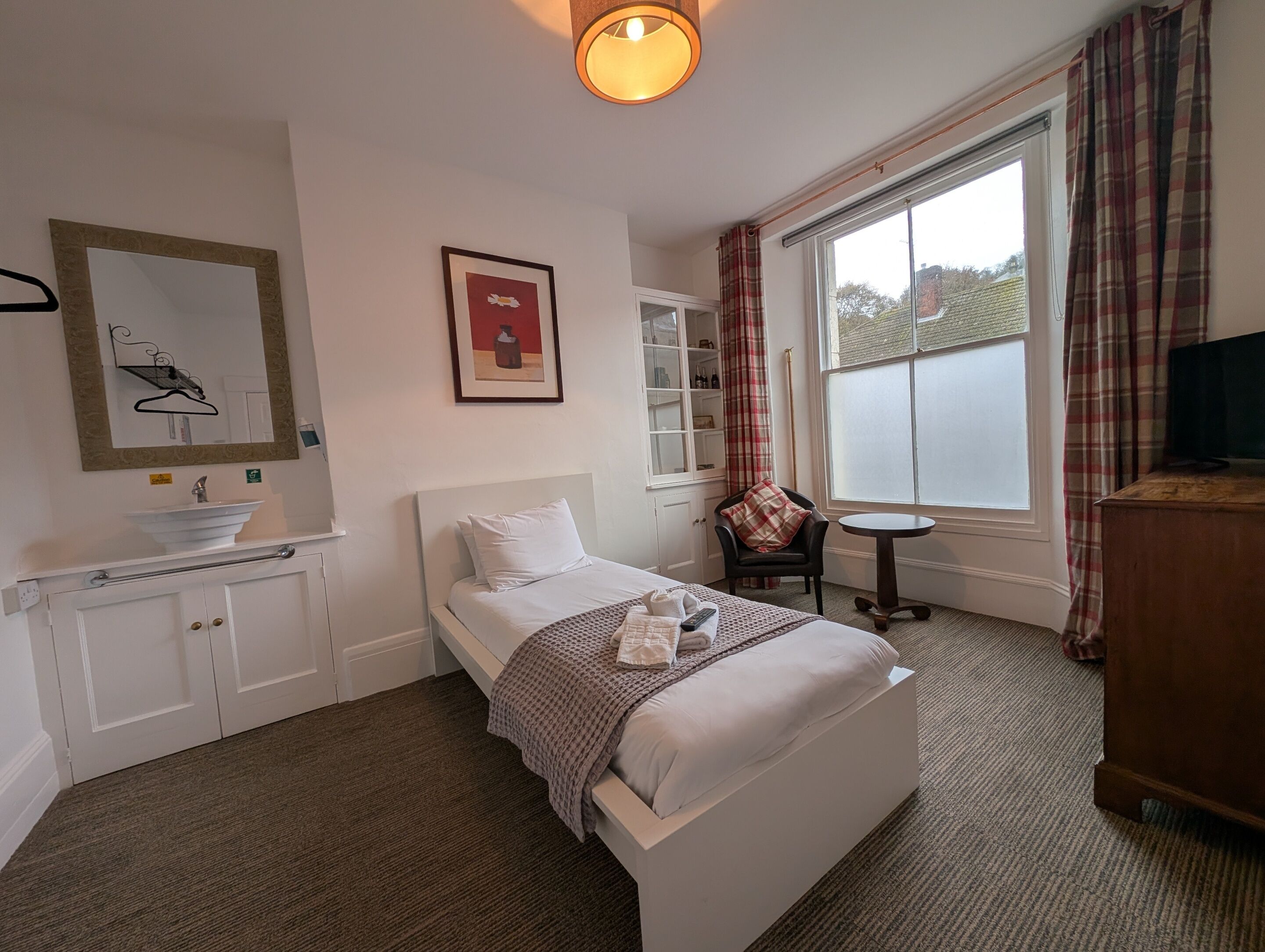 Habitación de hotel con cama, mesita de noche, una pequeña mesa redonda, una silla, ventana con cortinas, espejo, estante con botellas y televisión.