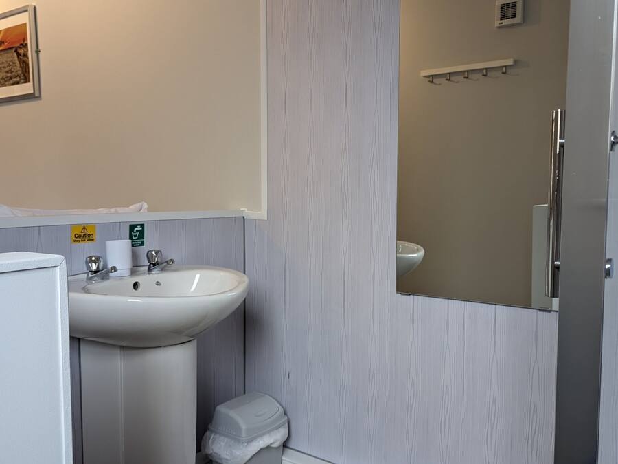 A bathroom with a sink, toilet, and shower. There is a towel dispenser and a caution sign on the wall.