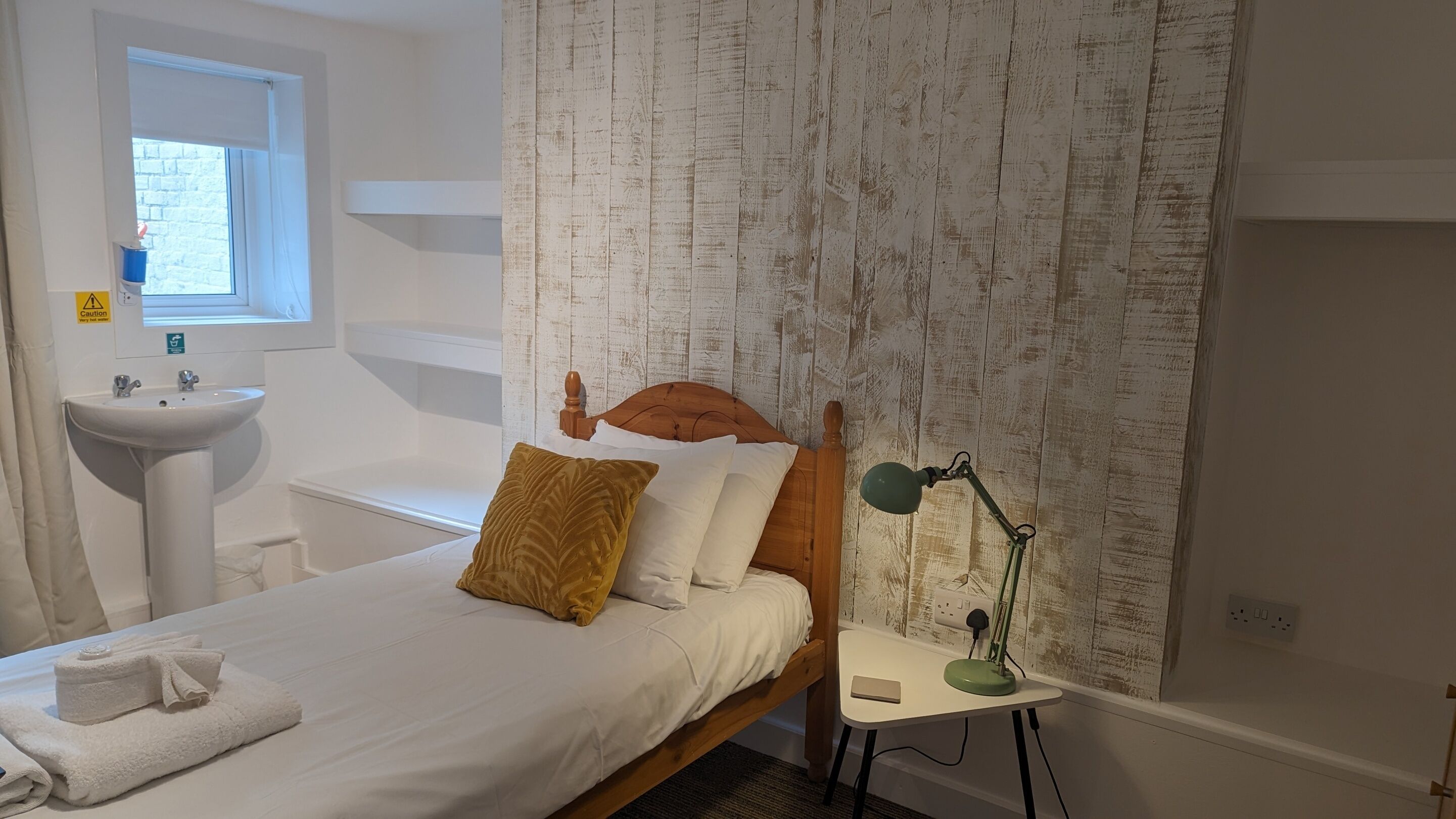 Ein Schlafzimmer mit einem Bett, einem Nachttisch mit Lampe, einem Fenster und einem Badezimmer mit Waschbecken.