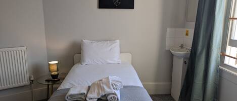 A hotel room with a bed, a bedside table with a lamp, a radiator, a sink, and a window with curtains.