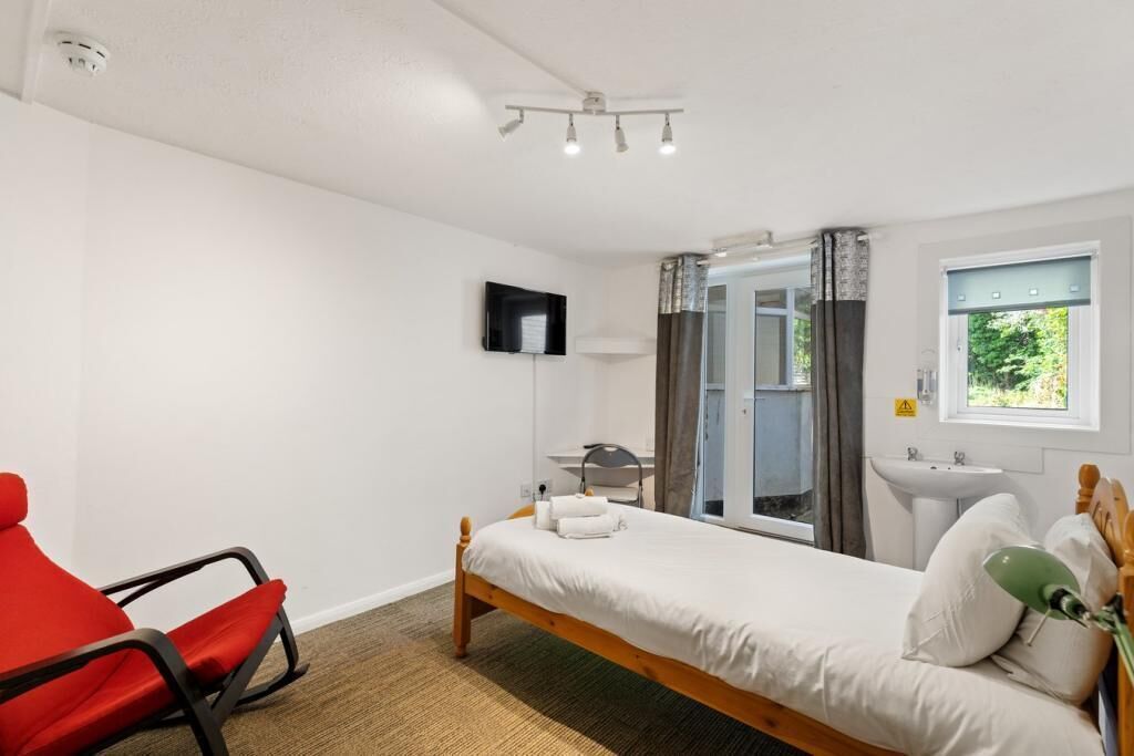 A bedroom with a bed, a red chair, a TV, a window, and a bathroom with a sink.