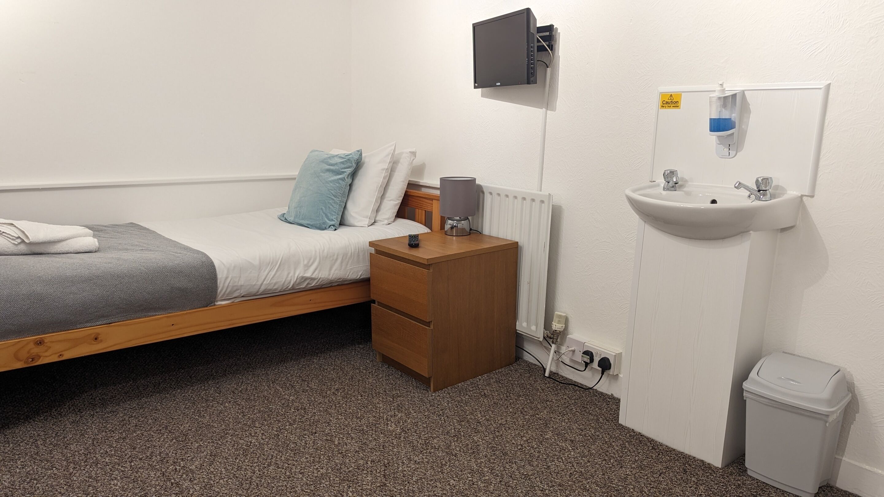 A small, tidy room with a single bed, a bedside table with a lamp, a wall-mounted TV, a white sink, and a trash bin.