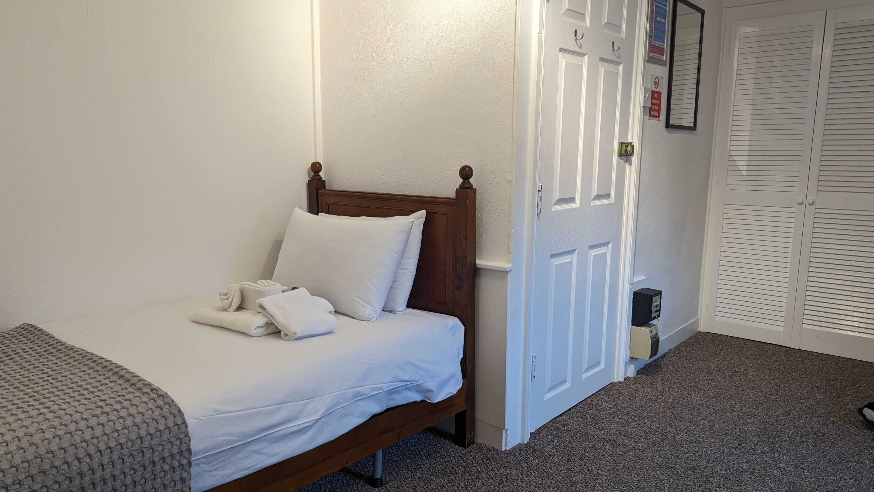 A neatly made bed with white linens and a wooden headboard, a small bedside table, and a closed white door with a sign prohibiting smoking.