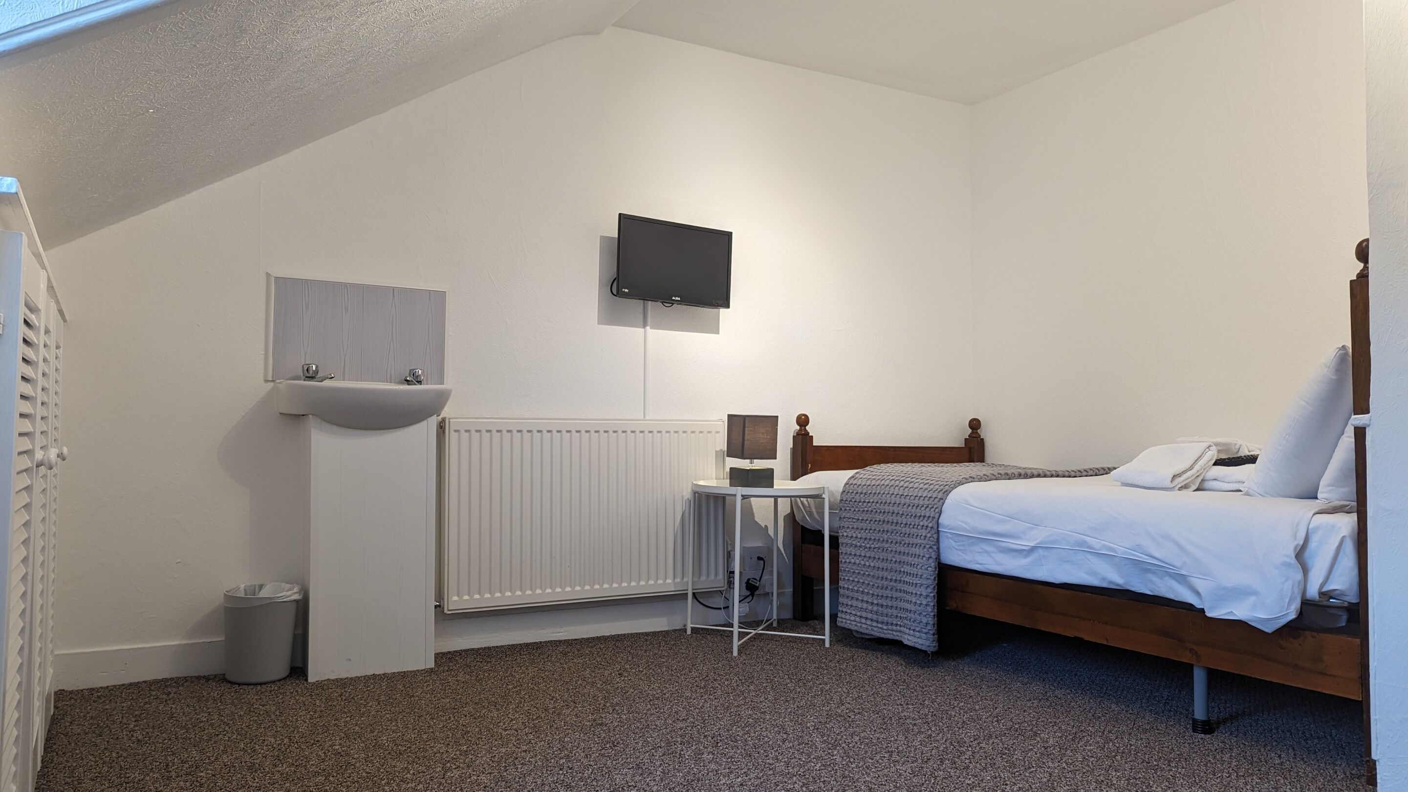 A bedroom with a single bed, a bedside table, a TV mounted on the wall, a white radiator, and a small window.