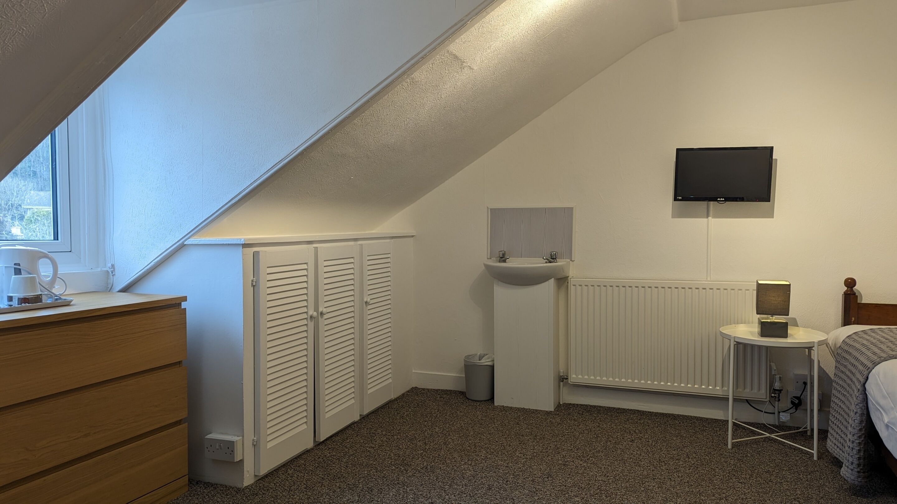 Une chambre avec un plafond en pente, un lit, une table de chevet avec une lampe, un radiateur, un téléviseur fixé au mur et une fenêtre.