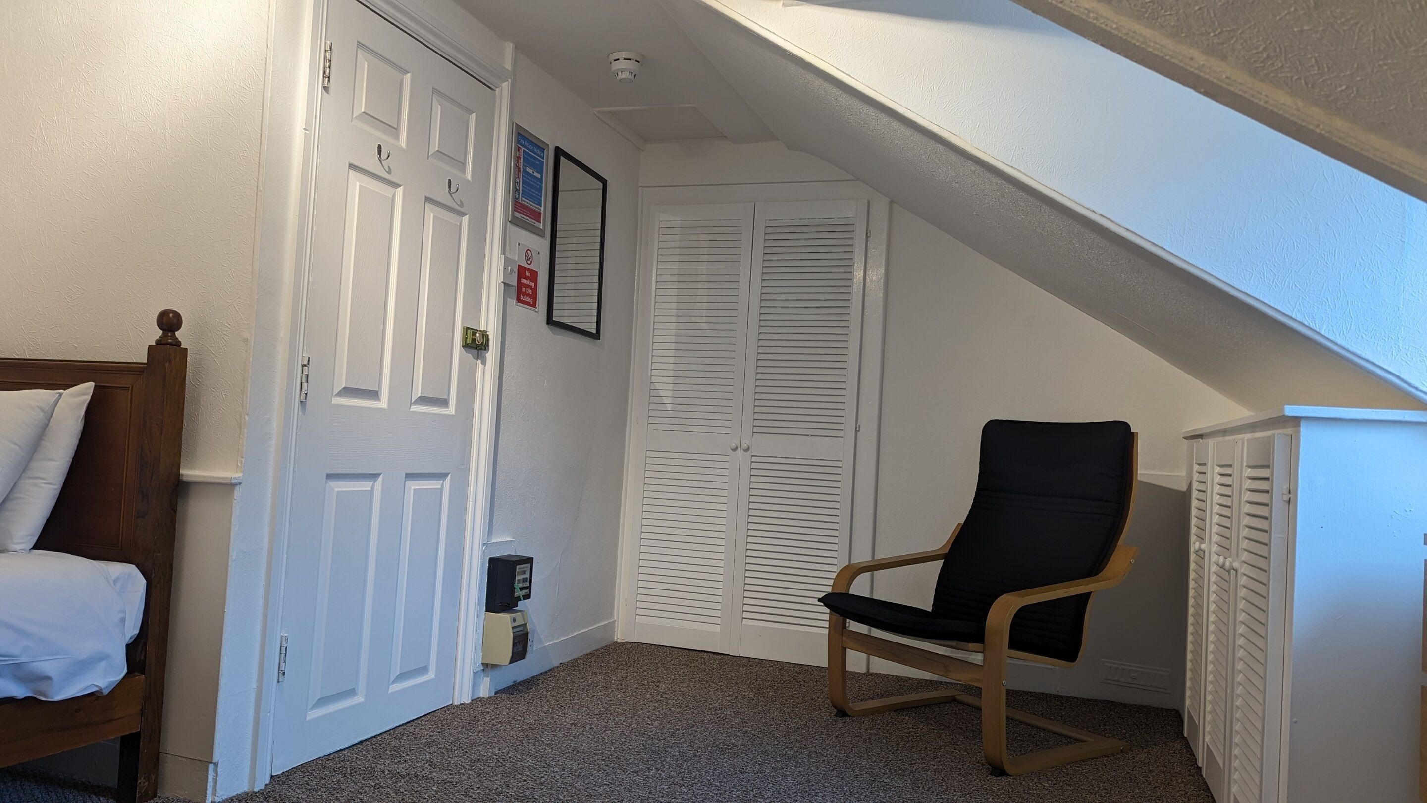 A bedroom with a bed, a chair, a closet, and a door with a smoke detector.