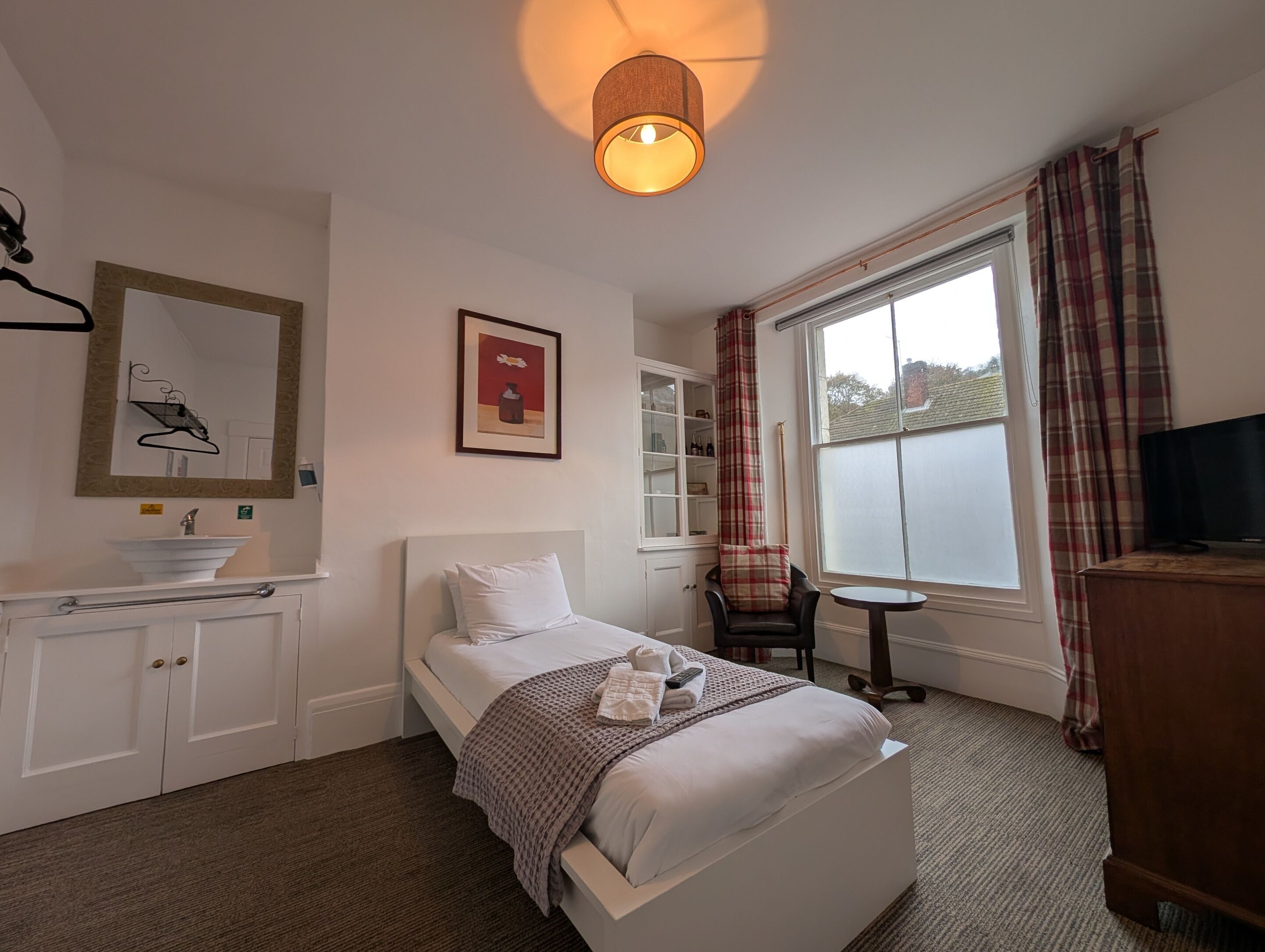 A hotel room with a bed, a bedside table, a chair, a TV, a window with curtains, a mirror, a cabinet, and a hanging light fixture.