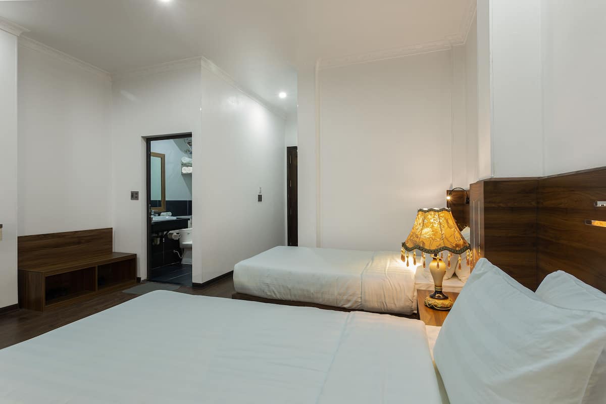 Une chambre d’hôtel avec deux lits, un banc en bois, une lampe de chevet et une salle de bain visible à travers une porte ouverte.