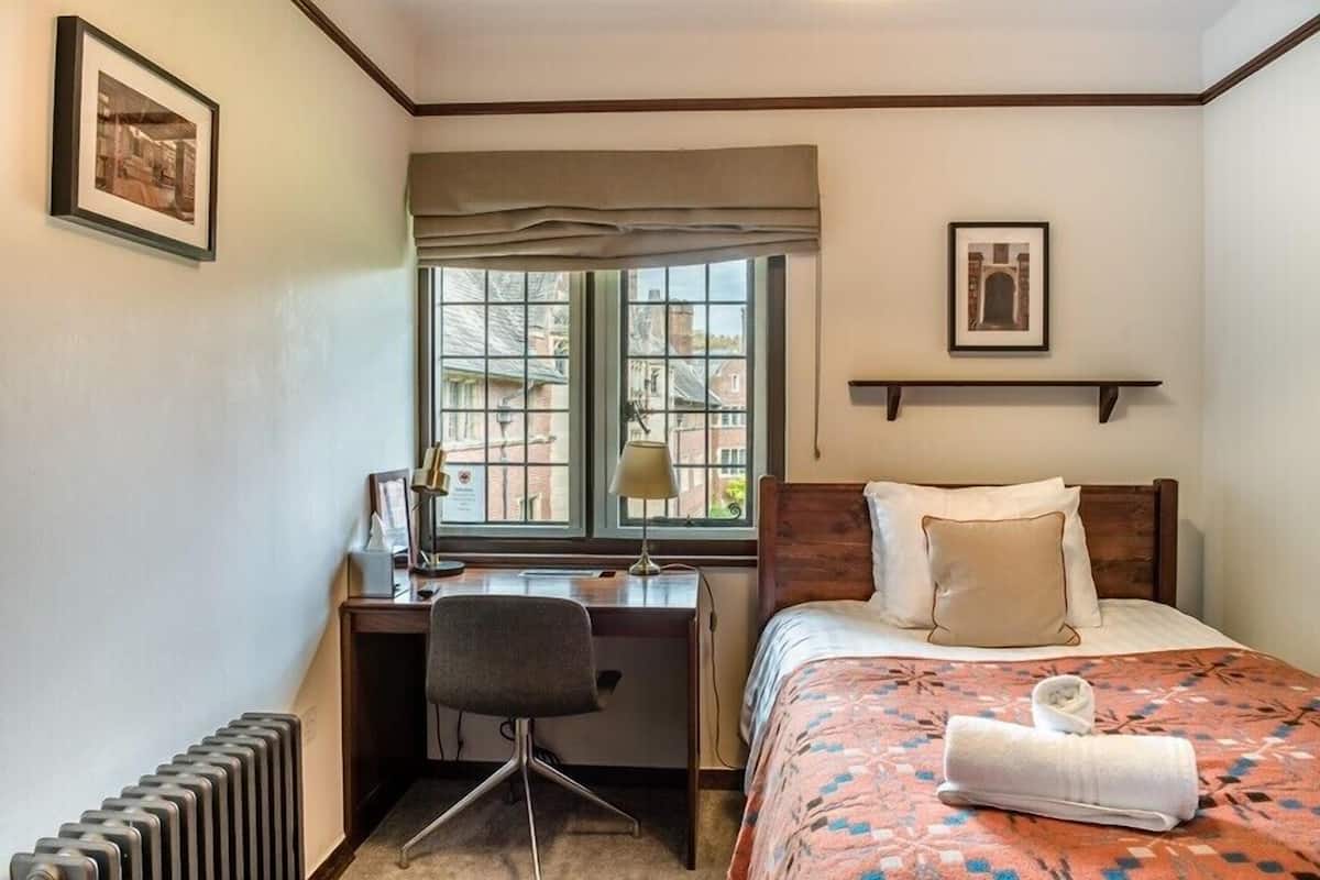 A hotel room with a bed, a desk, a chair, a radiator, a window with a view of buildings, and a framed picture on the wall.