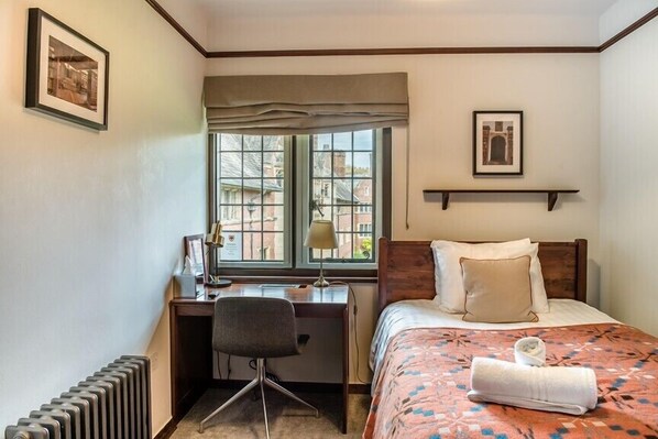 A hotel room with a bed, a desk, a chair, a radiator, a window with a view of buildings, and a framed picture on the wall. - West Court - Cambridge - Campus Accommodation (Cambridge)