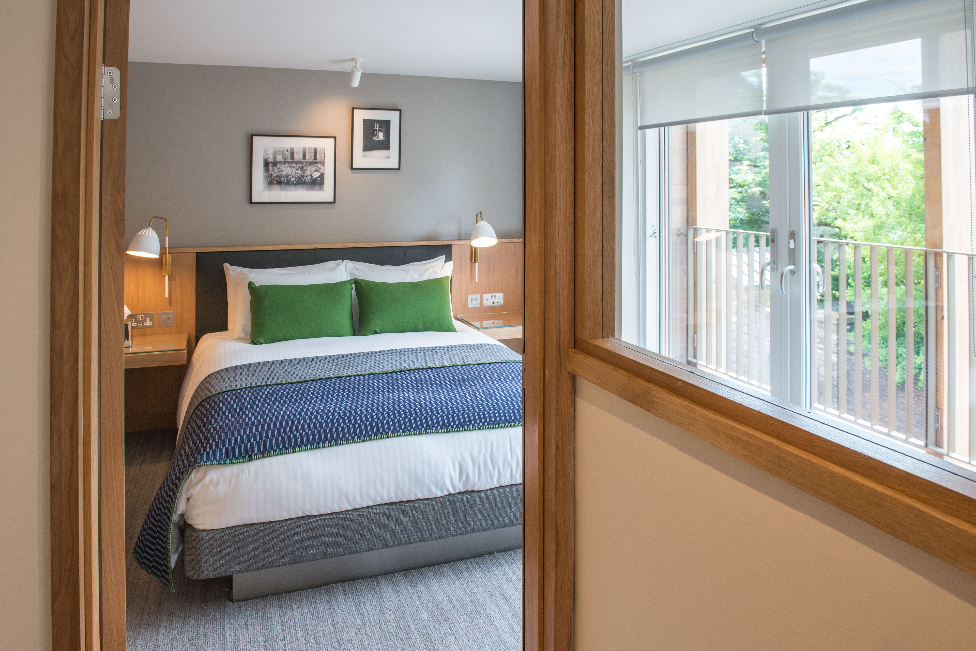A bedroom with a bed, two bedside lamps, a window with a view of greenery, and a framed picture on the wall.