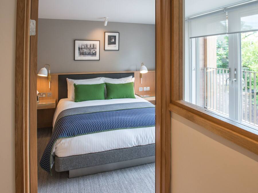 A bedroom with a bed, two bedside lamps, a window with a view of greenery, and a framed picture on the wall.