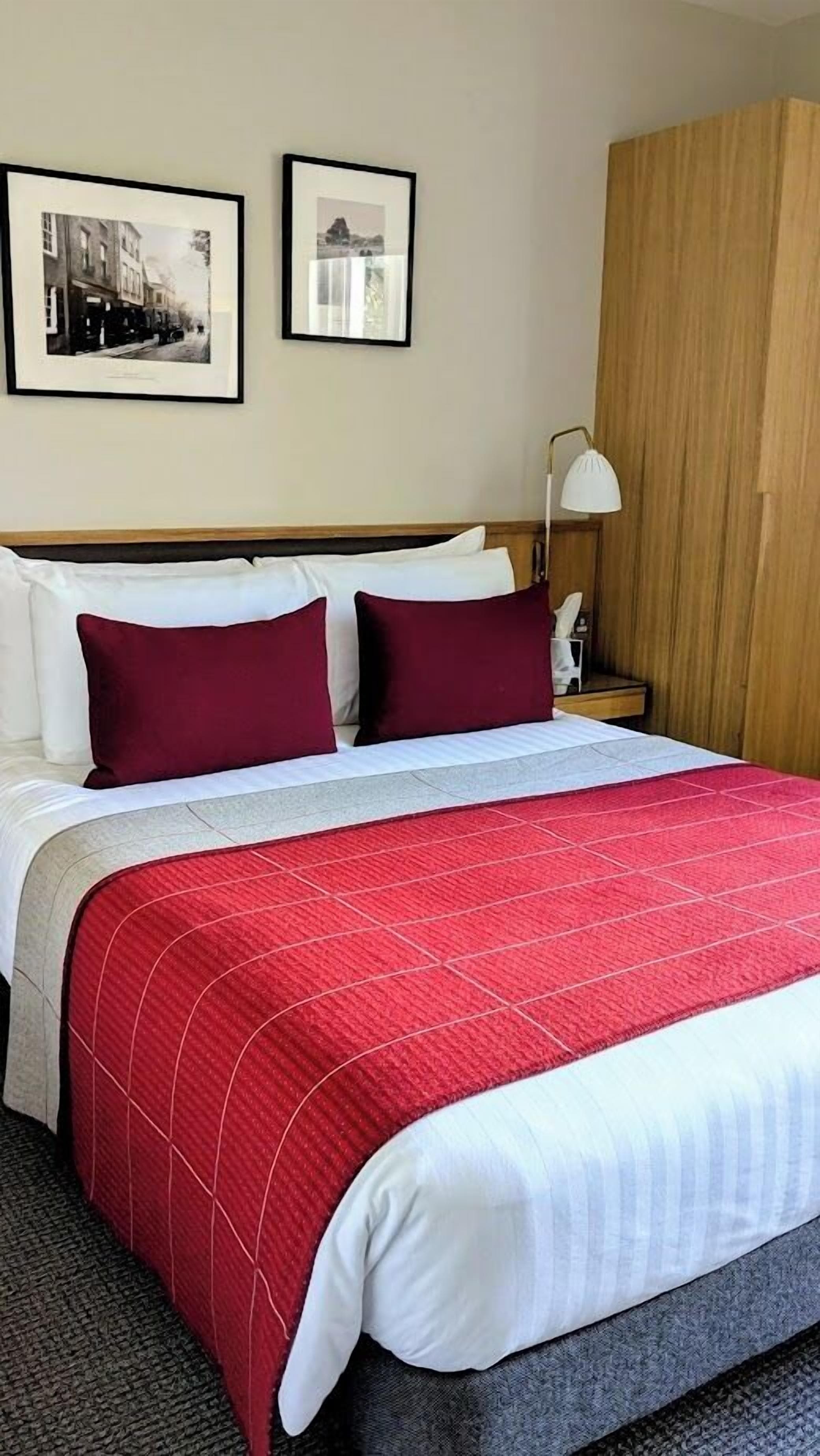 Una camera d'albergo con un letto, due quadri appesi al muro, un armadio in legno e un comodino con una lampada.