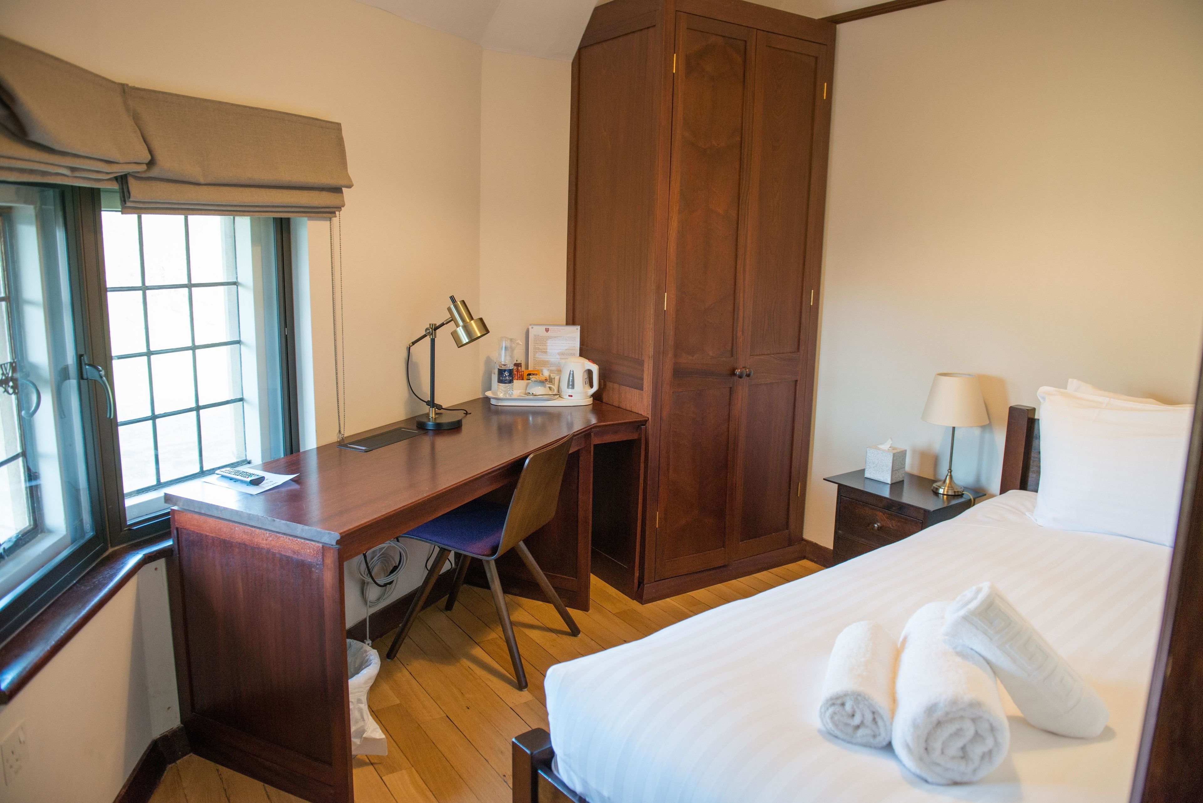 Een hotelkamer met een houten bureau, een stoel, een bed met witte lakens, een kledingkast, een nachtkastje en een raam met gordijn.