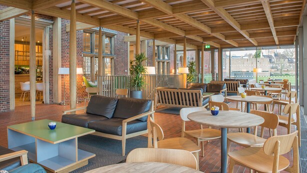 An indoor seating area with wooden tables and chairs, a sofa, and a brick wall. - West Court - Cambridge - Campus Accommodation (Cambridge)