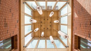 A modern interior with a central wooden ceiling structure, glass panels, and cylindrical light fixtures. - West Court - Cambridge - Campus Accommodation (Cambridge)