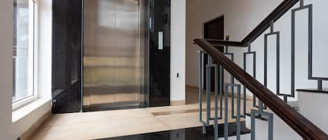 Un ascensor moderno con detalles metálicos, una escalera de madera con barandillas de metal y un suelo de tonos claros.