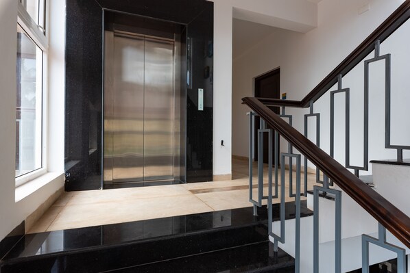 Un ascensor modern amb acabat metàl·lic, una escala de fusta amb barana de metall i un sòl de tons clars.