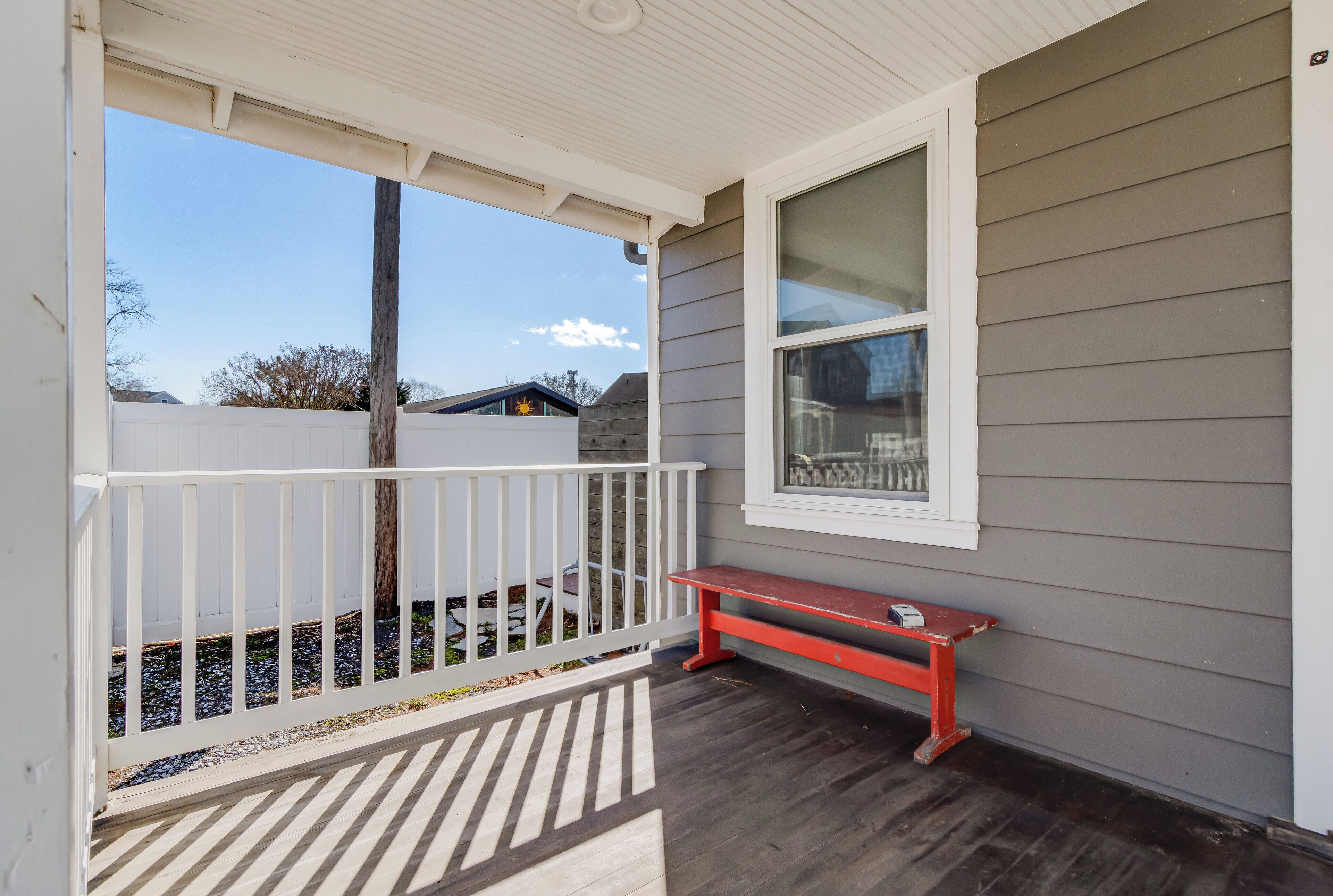 A covered porch with a red bench, a white railing, and a window overlooking a backyard.