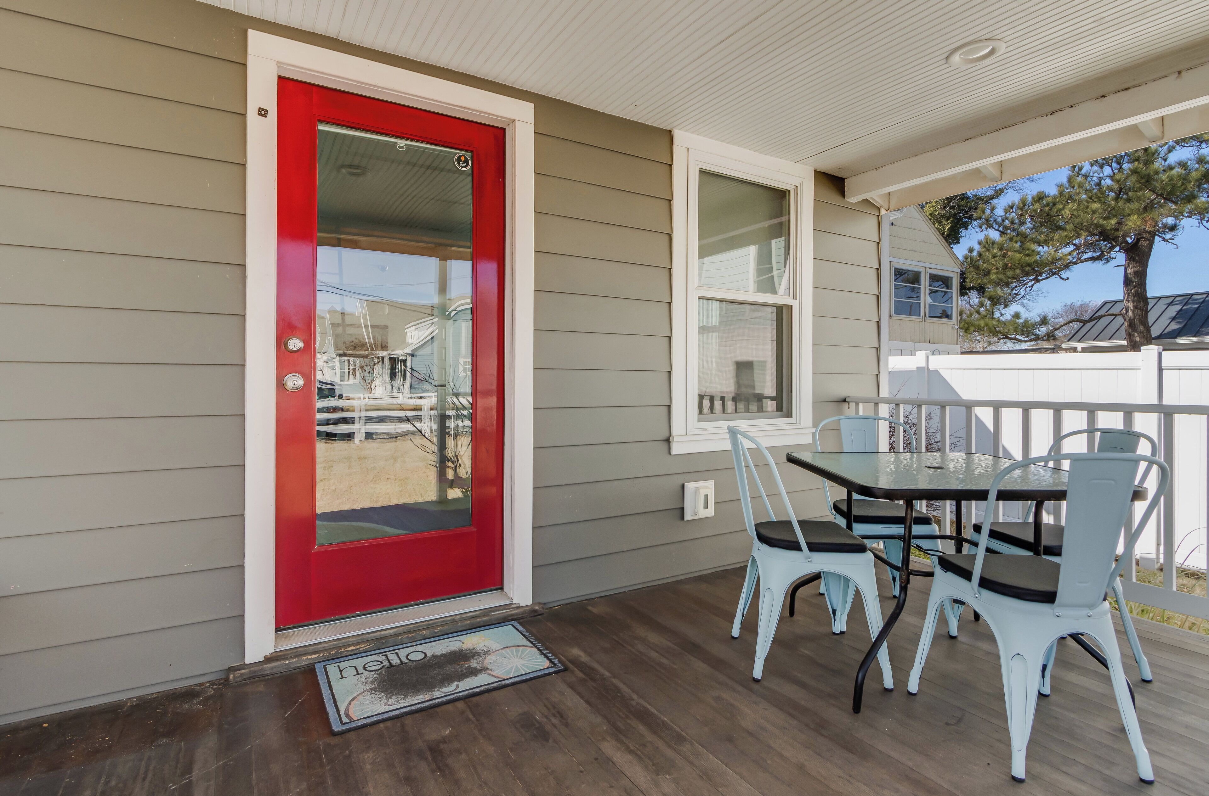 Un porche abrité avec une porte rouge, une table à manger avec des chaises et une vue sur la maison voisine.